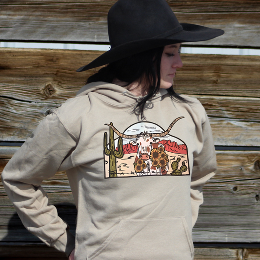 Cowgirl wearing khaki colored hoodie with longhorn cow scene with cactus and daisies. 