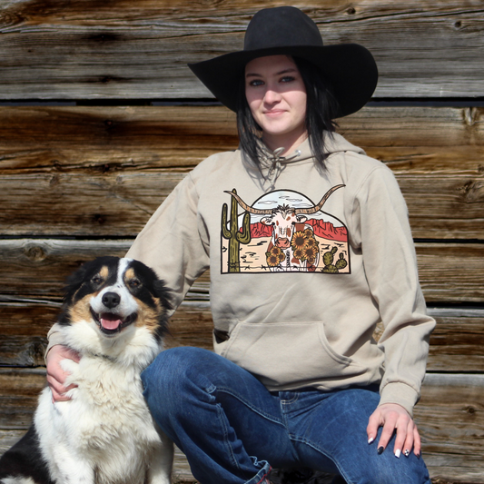 Cowgirl wearing khaki colored hoodie with longhorn cow scene with cactus and daisies. 